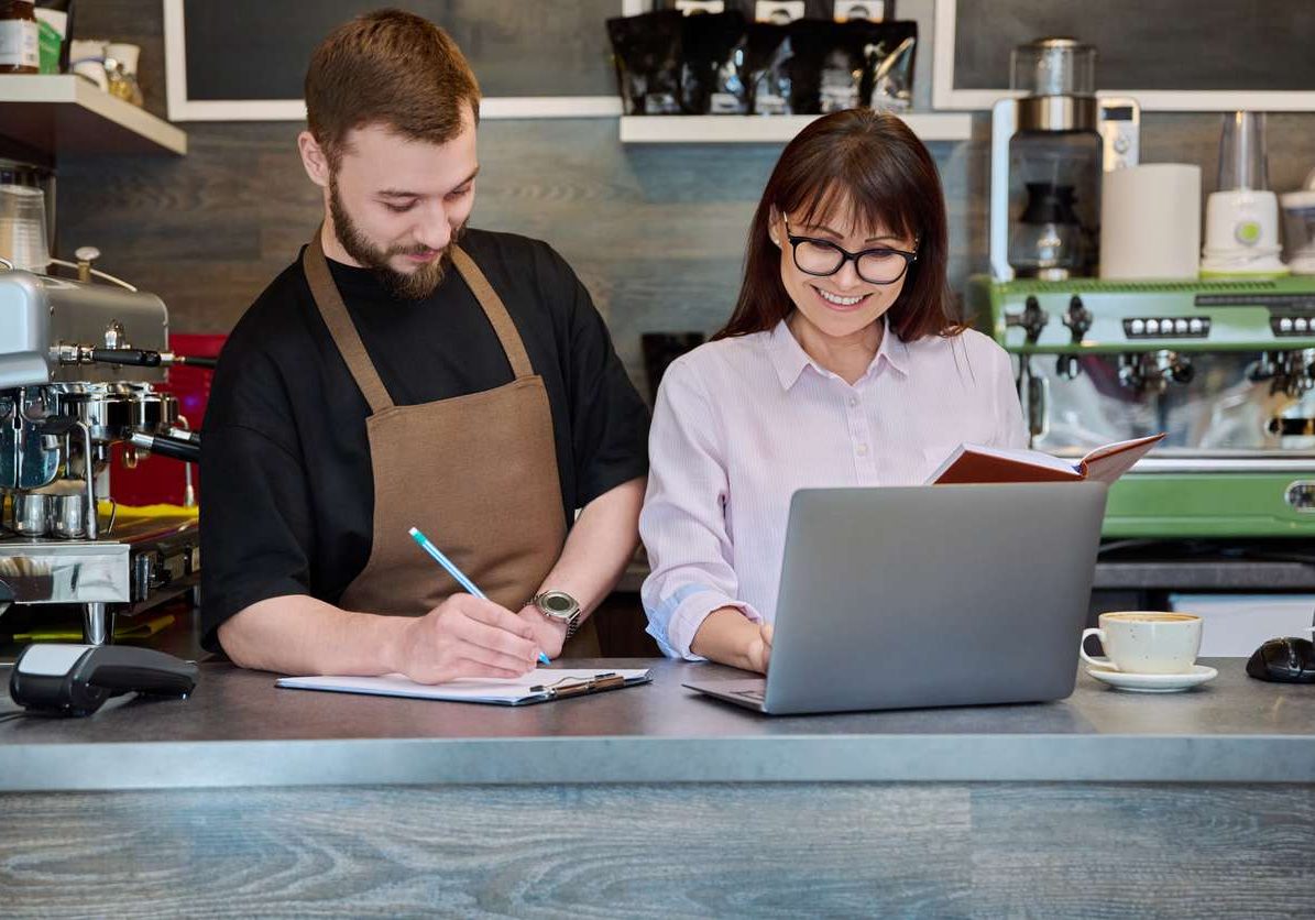 Colleagues partners, young male and mature woman talking working using laptop standing behind bar in coffee shop. Team, small business, work, staff, cafe cafeteria restaurant, entrepreneurship concept