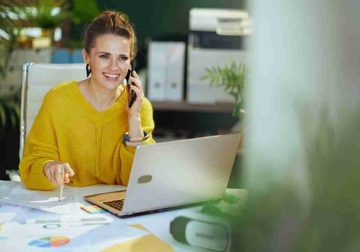 smiling trendy middle aged small business owner woman in yellow sweater with laptop speaking on a smartphone and working with documents in the modern green office.